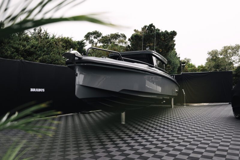 A sleek gray BRABUS luxury motorboat is displayed on stands atop a checkered platform, surrounded by black curtains and greenery—an impressive sight reminiscent of Pebble Beach’s celebrated cars.