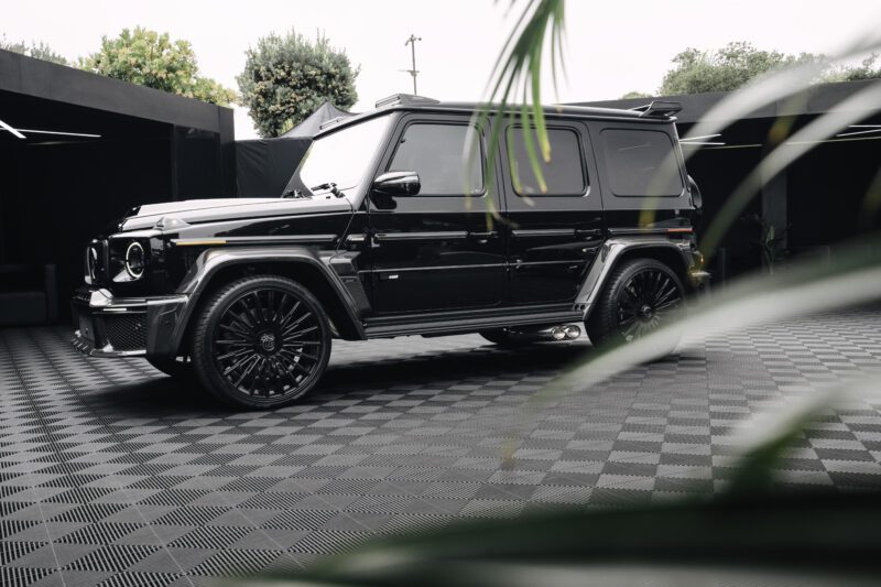 A black BRABUS Mercedes-Benz G-Class SUV is parked on a patterned floor at Pebble Beach, partially obscured by green leaves in the foreground.