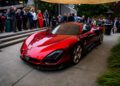 A red Alfa Romeo 33 Stradale supercar is displayed outdoors at an event, surrounded by a crowd of people on concrete steps and under a canopy.