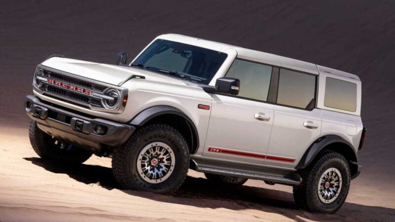 A white Ford Bronco SUV with off-road tires is parked on a sandy slope, showing its front and side profile—highlighting the rugged style celebrated as the Bronco turns 60: Ford reveals Heritage Edition for 2026.