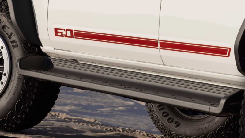Close-up of a white vehicle’s side displaying a red "6.0" decal and stripe, with black side step and rugged off-road tires—highlighting the Bronco Turns 60: Ford Reveals Heritage Edition for 2026.