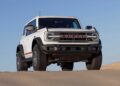 A white Ford Bronco Raptor SUV parked on sand dunes under a clear blue sky, celebrating Bronco Turns 60: Ford Reveals Heritage Edition for 2026.