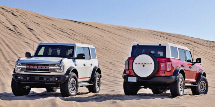 Two Ford Bronco SUVs, including a 2026 Ford Heritage Edition, are parked on sand dunes under a clear blue sky—one facing forward and the other backward.