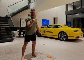 A man stands barefoot indoors in a ready stance for a Trick Shot Challenge, with a white dog nearby and a yellow Bentley Continental labeled “UNDERDOG” parked behind him.