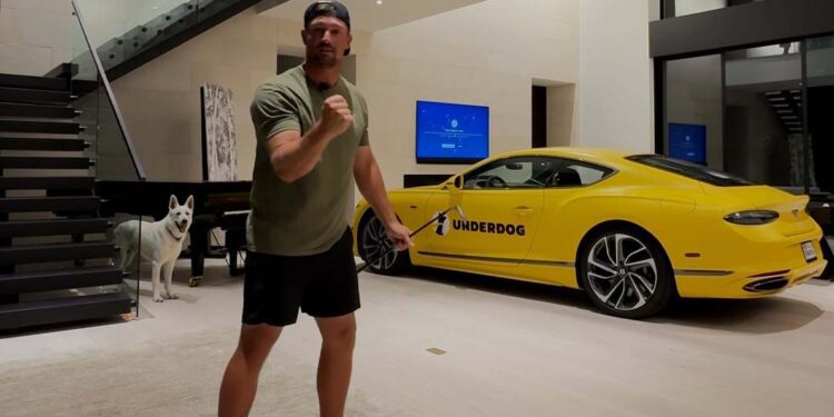 A man stands barefoot indoors in a ready stance for a Trick Shot Challenge, with a white dog nearby and a yellow Bentley Continental labeled “UNDERDOG” parked behind him.