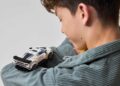 A boy in a gray jacket looks at a white toy car resting on his arm, as LEGO and Bugatti unite to celebrate the iconic Centodieci, set against a plain background.
