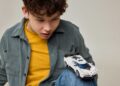 A boy in a yellow shirt and green jacket looks at a white toy car—where LEGO and Bugatti unite to celebrate the iconic Centodieci—balanced on his bent knee against a plain background.