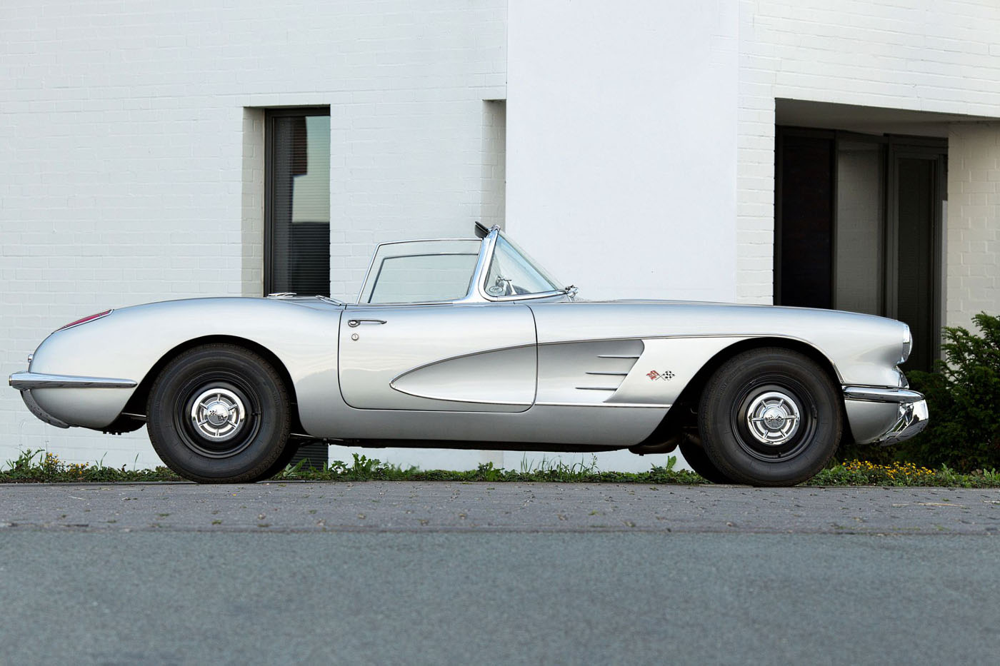 A Quail Silver Limited Edition Corvette ZR1X convertible is parked on a street in front of a white modern building, capturing the spirit of Monterey Car Week.