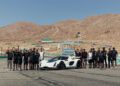 A group of people poses behind a white sports car on a race track at Willow Springs Raceway, with dry hills visible in the background.