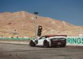 A white sports car with a red spoiler is parked on a racetrack near a traffic light, with desert hills and an American flag in the background.