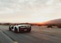 A white sports car with a large rear wing drives on an empty desert road at sunset, with mountains visible in the distance.