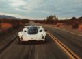 A white sports car drives down a deserted two-lane road through a desert landscape under a cloudy sky.