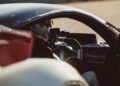 A person wearing a helmet sits in the driver's seat of a sports car, hands on the steering wheel, with sunlight shining through the windshield.