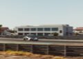 A white sports car driving at high speed on a racetrack with blurred motion, modern buildings, and barriers in the background.