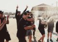 A group of people in black shirts celebrate outdoors, spraying champagne and cheering on a sunny day at what appears to be a racetrack.