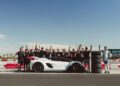 A group of people stands behind a white sports car on a racetrack, holding up one finger; a person in a racing suit stands by a stack of tires on the right.