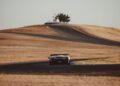 A sports car drives on a winding, paved race track surrounded by dry grass and rolling hills under a clear sky.