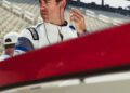 A man in a racing suit adjusts his earpiece next to a red and white race car at a racetrack, with empty grandstand seats in the background.