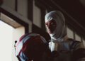 A racecar driver in a white suit and balaclava stands indoors, looking down at a red and white helmet in his hands.
