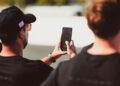 Two people stand outdoors; one holds up a smartphone displaying a timer at 1:35. The scene appears to be in daylight near a track or roadway.