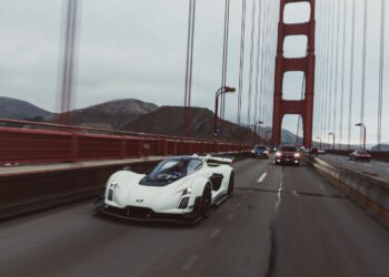 A white sports car drives on a bridge alongside other vehicles under an overcast sky, with suspension cables and hills visible in the background.
