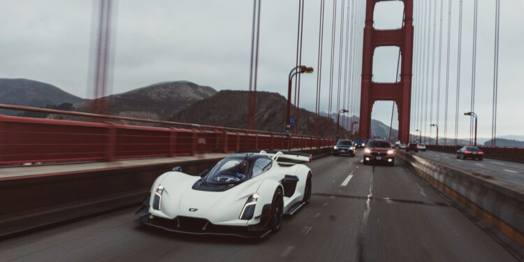 A white sports car drives on a bridge alongside other vehicles under an overcast sky, with suspension cables and hills visible in the background.