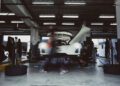 A white sports car with open doors is being serviced in a garage, with tires and equipment around and people working on or near the vehicle.