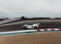 A white sports car speeds around a racetrack curve, with blurred background and motion indicating high velocity.