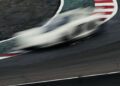 A blurred white race car speeds around a sharp curve on a racetrack with red and white striped curbing.