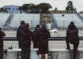 A group of people in matching black jackets stand by a racetrack, observing a white race car driving past empty bleachers.