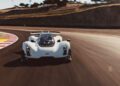 A white, futuristic sports car speeds around a racetrack curve, with blurred asphalt and red-and-white curbs visible in the background.