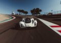 A white race car drives on a curved racetrack with blurred motion, passing by trees, a fence, and a small structure under a clear sky.