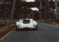 A white sports car speeds down a narrow, tree-lined road with a blurred background, indicating motion.