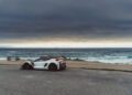 A white sports car is parked near a coastal road with the ocean and a cloudy sky in the background.