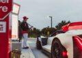 A person in uniform takes an order from the driver of a white sports car at a fast-food drive-thru.