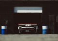 A white sports car with a red rear wing is parked inside a dark garage, partially visible under a half-open door, with blue and white barrels on each side.