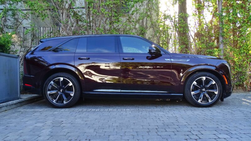 A dark purple Cadillac Escalade IQ, an all-electric SUV, is parked on a stone driveway beside a wall and trees with green foliage in the background.