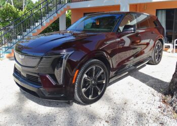 A dark maroon Cadillac Escalade IQ, a luxury all-electric SUV, is parked on a gravel driveway in front of an orange building with white trim and a staircase.