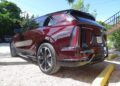 A maroon 2025 Cadillac Escalade IQ Sport 2, a gigantic luxury all-electric SUV that can drive itself, is parked on a gravel surface near a yellow curb, next to some trees and a stairway.