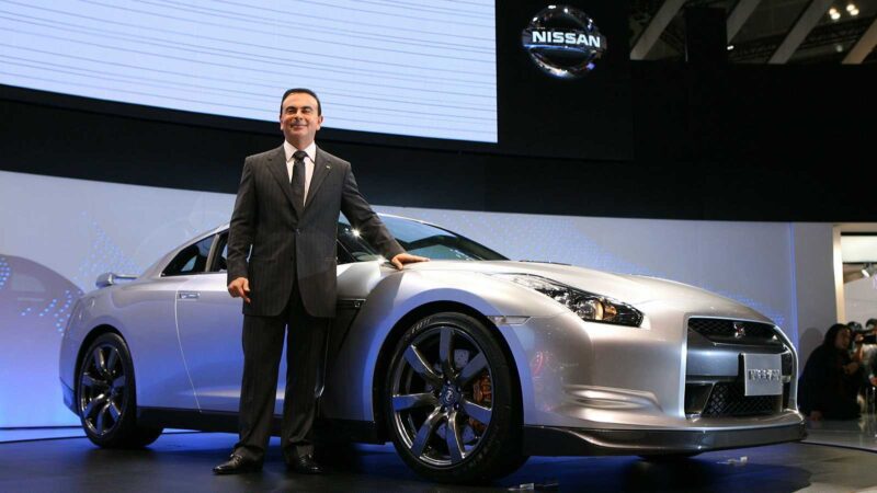 A man in a suit stands next to a silver Nissan R35 GT-R on display at an auto show, highlighting the next generation of performance, with a Nissan logo visible in the background.