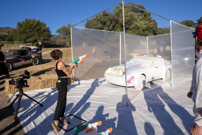 A person sprays colored liquid onto a white car with a water gun inside a transparent enclosure, while several people watch outdoors during Car Week.