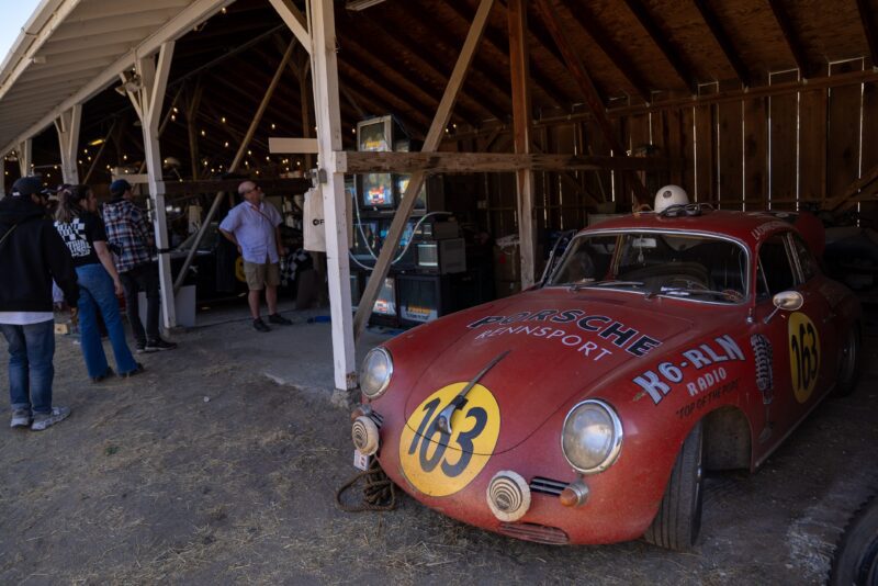 A vintage red Porsche race car with number 163 is parked outside a wooden shed, where several people stand near arcade machines—an Off the Beaten Path gem for Car Week enthusiasts.