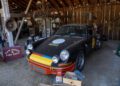 A black vintage Porsche with racing decals is parked inside a rustic garage filled with automotive tools, signs, and equipment—a true Sportscar Vacationland for enthusiasts off the beaten path.
