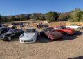 A row of classic and vintage sports cars parked on a dirt lot during Car Week, with people strolling around, trees and hills in the background under a clear blue sky—perfect for those seeking an off the beaten path sportscar vacation.