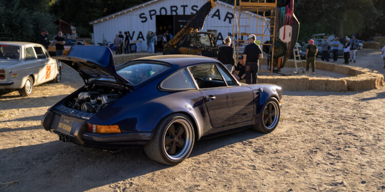 A blue classic Porsche with its rear trunk open is parked on a dirt lot near a white building labeled "SPORTS CAR"—an ideal Off the Beaten Path stop during your Car Week Escape, with people and other vehicles in the background.