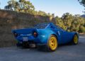 A blue vintage sports car with yellow wheels is parked on a dirt road beside stacked hay bales and trees—perfect for an off the beaten path sportscar vacation.