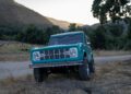 A teal vintage Ford Bronco is parked on a dirt road, off the beaten path, with trees and mountains in the background at sunset.