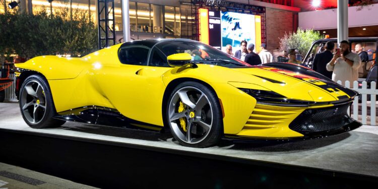 A bright yellow sports car, possibly one of the 10 Most Expensive Cars To Sell At 2025 Monterey Car Week Auctions, is displayed on a platform outdoors at night, with people and a building visible in the background.