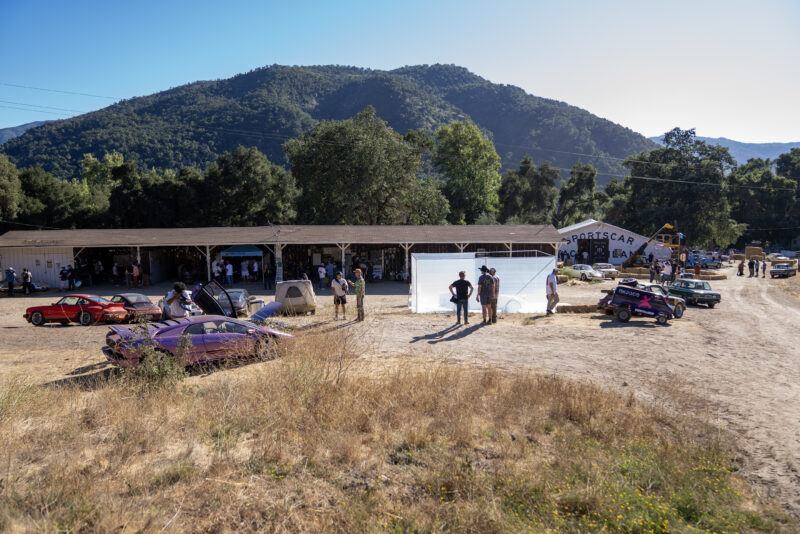 A rural outdoor car event, part of Car Week Escape, with people gathered near a building, several parked cars, a white tent, and wooded hills in the background.
