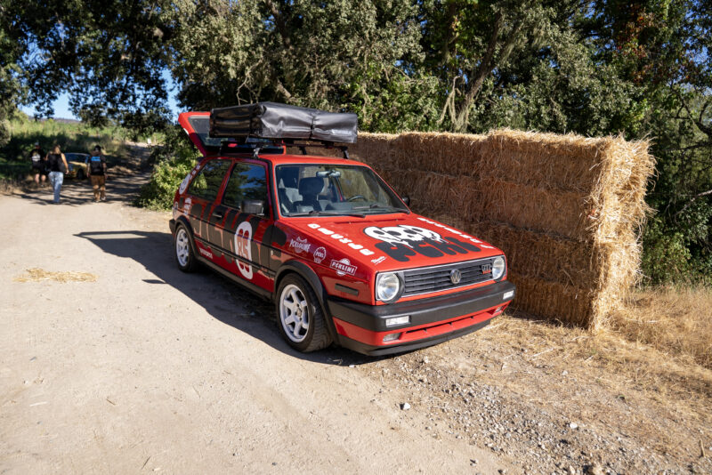 A red Volkswagen Golf GTI with custom decals and a roof box is parked on a dirt road beside stacked hay bales and trees, capturing the spirit of Sportscar Vacationland for those seeking adventures off the beaten path.